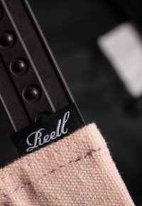 Pink fabric tag with textured surface and black lettering labeled "Reell," attached to a dark strap featuring blunt ridges.
