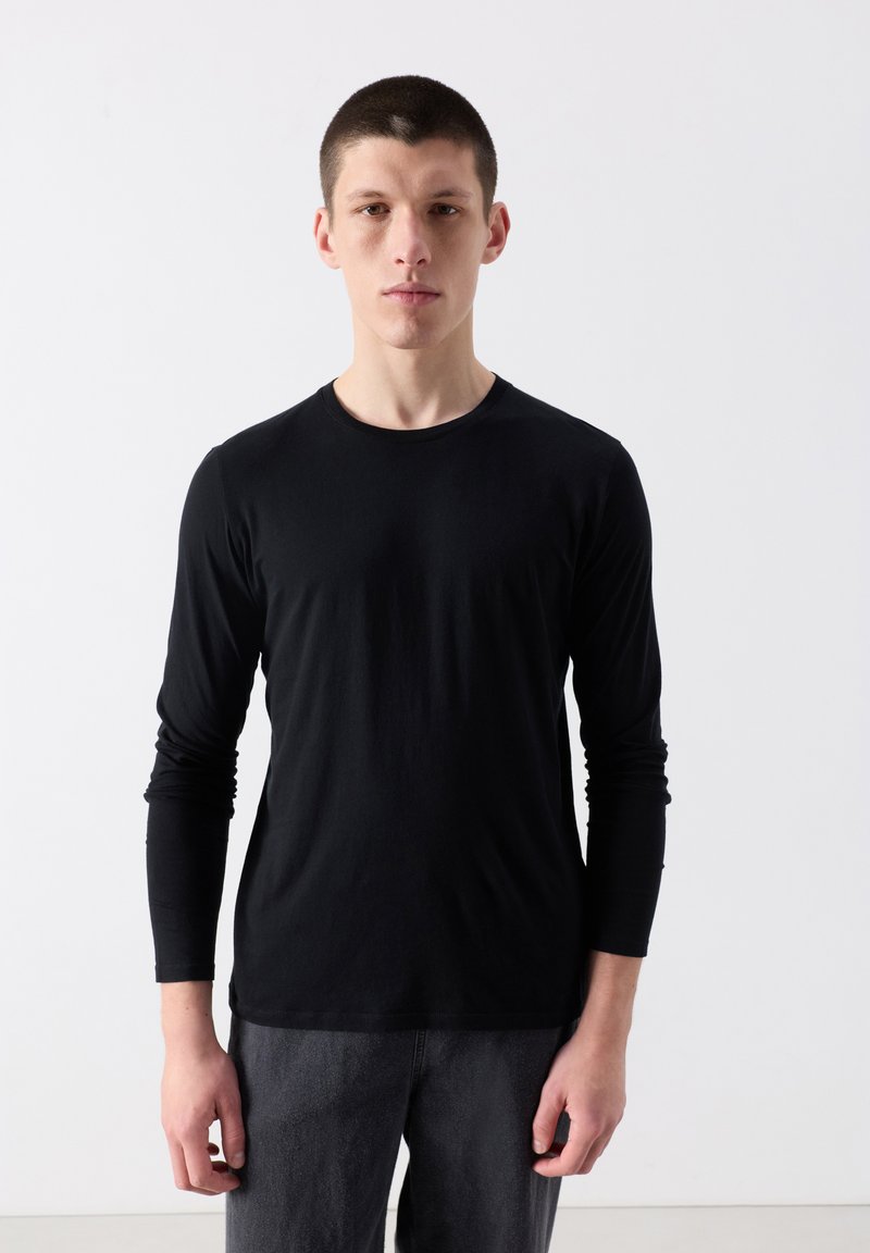 Noor mees lühikeste juustega, musta pika varrukaga särgi ja tumedate pükstega, seismas lihtsa valge tausta ees.