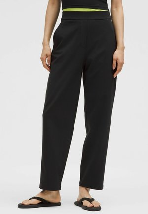 Schwarze, weit geschnittene Hose mit glatter Textur, hoher Taille und diskret eingearbeitetem Fronttasche, kombiniert mit schwarzen Flip-Flops.