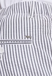 Proužkované šedé a bílé kalhoty s knoflíkovým zapínáním a malou kapsou s kovovou značkou značky. Měkká tkanina s texturovaným povrchem.