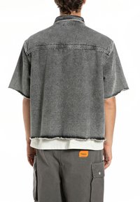 Camicia in denim grigia a maniche corte con bordi grezzi e colletto ripiegato. Indossata sopra una camicia bianca, abbinata a pantaloni cargo grigi.