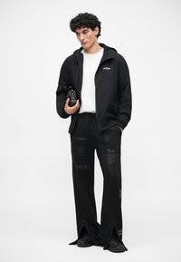 Melnā jaciņa ar rāvējslēdzēju virs balta krekla, kombinēta ar augsto vidukli melnām biksēm, kurām ir dažādi raksti un akcenti, uz balta fona.