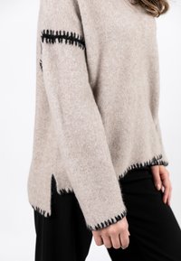 Beige Wollpullover mit kontrastierendem schwarzen Stich entlang der Kanten und Ärmel, der eine lockere Passform und seitliche Schlitze am Saum aufweist.