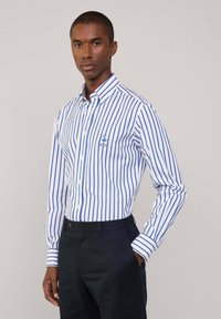 Camisa blanca con rayas verticales azules, diseño de botones, detalle de bolsillo de marca, mangas largas, combinada con pantalones oscuros.