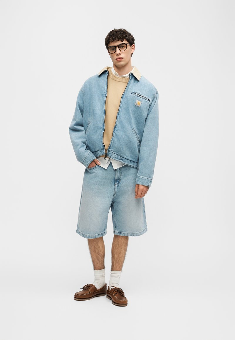 Jonge man met een lichtblauwe spijkerjas en korte broek, beige trui, witte sokken, bruine schoenen en een zwarte bril tegen een effen achtergrond.