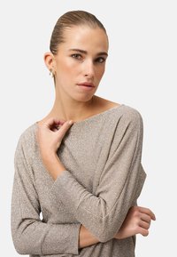 Frau mit zurückgekämmtem Haar, die goldene Ohrringe und ein funkelndes Langarmoberteil trägt und mit einer Hand den Ausschnitt hält.