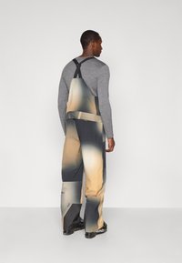 Άνδρας με γκρι μακρυμάνικο πουκάμισο και σορτσάκι με gradient σχέδιο, φορώντας μαύρες μπότες σκι, να στέκεται σε απλό φωτεινό γκρι φόντο.