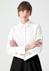 Camisa blanca de botones, hecha de tela suave, con un cuello estructurado y puños anchos con acentos de botones dorados, combinada con una falda negra.