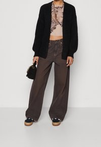Černý chlupatý cardigan přes vzorovaný crop top, spárovaný s volnými hnědými džíny a platformovými černými botami, držící malou černou kabelku.