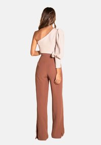 Top asimétrico en beige claro con una manga abullonada, combinado con pantalones marrones de cintura alta que tienen aberturas laterales. Textura suave en toda la prenda.