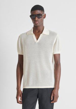 Jeune homme portant des lunettes de soleil noires, un polo en maille blanc à manches courtes et un pantalon gris foncé, debout devant un fond clair uni.