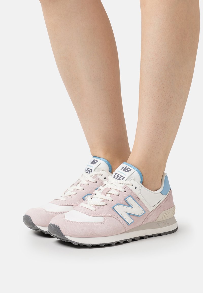 Pink and white sneakers featuring suede and mesh, with blue accents and a black rubber sole. Model 574 displayed on tongue.