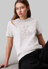 Joven mujer con cabello largo que lleva una camiseta gris claro de Calvin Klein Jeans y pantalones negros, sentada contra un fondo gris liso.