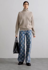 Beiger gerippter Rollkragenpullover, kombiniert mit weit geschnittenen Jeans in Blau mit Sternenmuster und einer schwarzen Tragetasche. Glatte, texturierte Materialien.