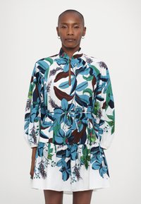 Blumenkleid mit einer Mischung aus blauen, grünen und braunen Blättern auf weißem Hintergrund, mit gerafftem Taillenzug und langen, gepufften Ärmeln.