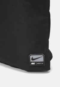 Sort Nike Utility 2.0 taske med et tekstureret stof, der har et rektangulært logo patch i grå med et prikkemønster og sort skrift.