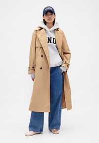 Beżowy płaszcz trencz z podwójnym rzędem guzików, zestawiony z szarą bluzą z kapturem ozdobioną czarnym nadrukiem. Noszony z szerokimi dżinsami o kroju wide-leg w kolorze niebieskim i beżowymi butami.