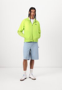 Neon green zip-up lightweight jacket with two front pockets, paired with light blue patterned shorts and white sneakers with beige accents.