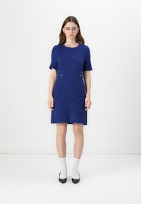 Blauwe gebreide jurk met korte mouwen, met een gestructureerd patroon en twee metalen accenten bij de taille. Het model draagt witte sokken en zwarte hakken.