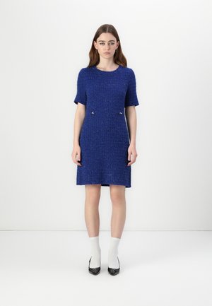 Blauwe gebreide jurk met korte mouwen, met een gestructureerd patroon en twee metalen accenten bij de taille. Het model draagt witte sokken en zwarte hakken.