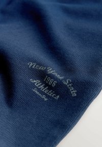 Nærbillede af mørkeblå strikket stof med hvidt trykt tekst, herunder "1986" og "Premium", på en hvid baggrund.