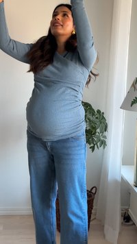 Zwangere vrouw in een blauw shirt en spijkerbroek die binnenshuis omhoog reikt, vlakbij een plant en een lamp met witte gordijnen op de achtergrond.