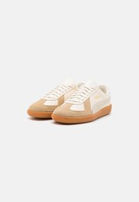 Zapatillas deportivas en tonos beige y crema, confeccionadas con materiales de ante y cuero, presentan una suela de goma, cordones y un detalle de logo en el lateral.