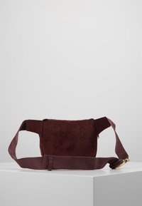 Ciemnobrązowa torba na biodra z korduroyu z regulowanym paskiem i złotą klamrą, wyeksponowana na białym pedestal na jednolitym szarym tle.