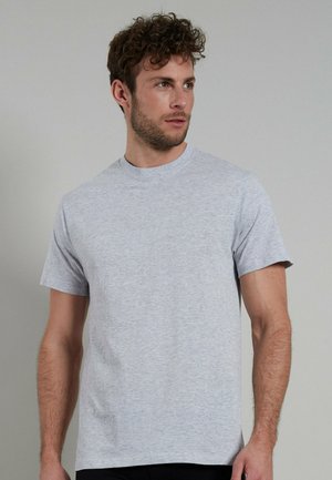 Mann mit lockigem Haar, der ein einfaches hellgraues T-Shirt mit kurzen Ärmeln trägt, schaut nach rechts und steht vor einem einfarbigen grauen Hintergrund.