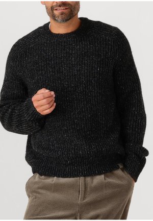 Mann in einem groben schwarzen Strickpullover und beigen Cordhosen, mit einer Hand teilweise in der Tasche und der anderen leicht erhoben.