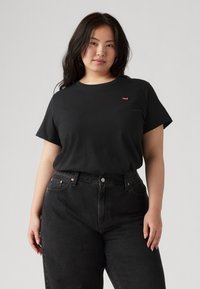 Black cotton t-shirt with short sleeves, round neckline, and a small red logo on the chest. Paired with high-waisted black jeans.
