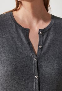 Zbliżenie na osobę noszącą ciemnoszary sweter z dzianiny z błyszczącymi srebrnymi guzikami, z których trzy są zapięte w okolicy klatki piersiowej.