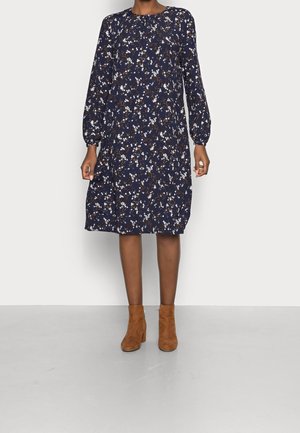 Femme portant une robe bleu marine longueur genou à imprimé floral blanc et manches longues bouffantes, associée à des bottines en daim marron.