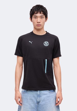 Jeune homme portant un t-shirt Puma noir avec un logo bleu clair et un détail de fermeture éclair, associé à un jean denim bleu clair, debout devant un fond uni.