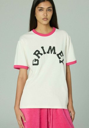 Giovane donna con capelli scuri e lisci che indossa una T-shirt bianca con rifiniture rosa e la scritta "GRIMEY", abbinata a pantaloni rosa acceso con texture.