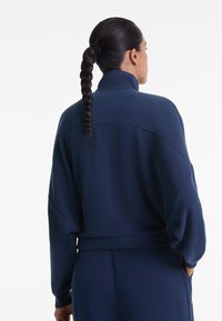 Marineblaue Sportjacke mit hohem Kragen, lockerem Schnitt und überschnittenen Schultern. Sie zeichnet sich durch eine glatte Textur und minimale Nähdetails aus.