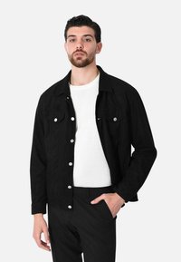 Giacca di jeans nera con due tasche frontali, bottoni a pressione argentati e colletto. Indossata sopra una camicia bianca, abbinata a pantaloni neri.