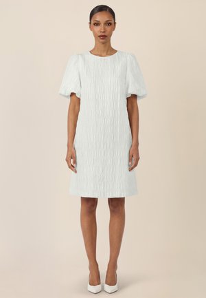 Frau steht und trägt ein weißes, strukturiertes, knieumspielendes Kleid mit kurzen Puffärmeln und weiße spitze Schuhe vor einem beigen Hintergrund.