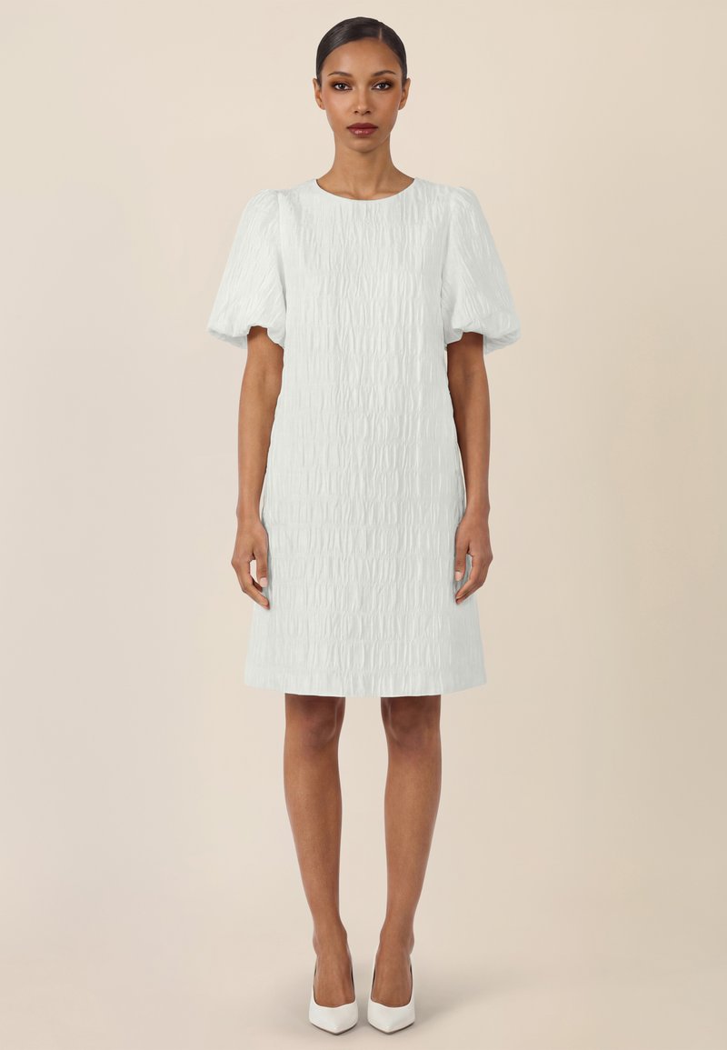 Femme debout portant une robe blanche texturée mi-longue avec des manches courtes bouffantes et des talons blancs à bouts pointus, sur un fond beige.