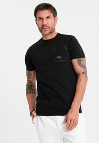 Svart bomullströja med rundad hals, en framficka med en logotyp och en slim-fit design. Tyget verkar mjukt och lätt.