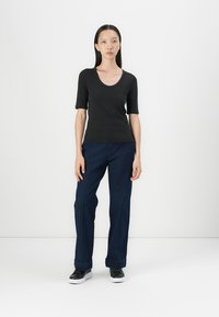 Zwarte korte mouwen shirt gecombineerd met blauwe denim broek met stiksel-details. Model draagt zwart-witte sneakers. Minimalistische achtergrond.