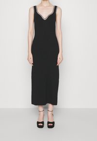 Robe en tricot noire avec un décolleté en V profond, texture côtelée et bordure décorative. Portée avec des talons compensés noirs à brides de cheville.