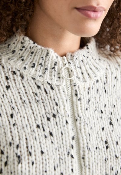 Strickjacke mit Reißverschluss in Creme mit schwarzen Flecken. Verfügt über einen hohen Kragen und strukturiertes Material, das horizontale Rippungen und einen Metallreißverschluss zeigt.