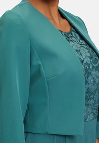 Tyrkysový blazer se štíhlým designem a zkráceným střihem, s hladkým materiálem a krajkovým detailem uvnitř v odpovídající barvě.