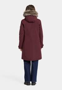 Burgundy parka dengan tudung bulu tiruan, reka bentuk hingga ke lutut, tekstur yang halus, dan poket sisi, dipadankan dengan seluar denim biru gelap.