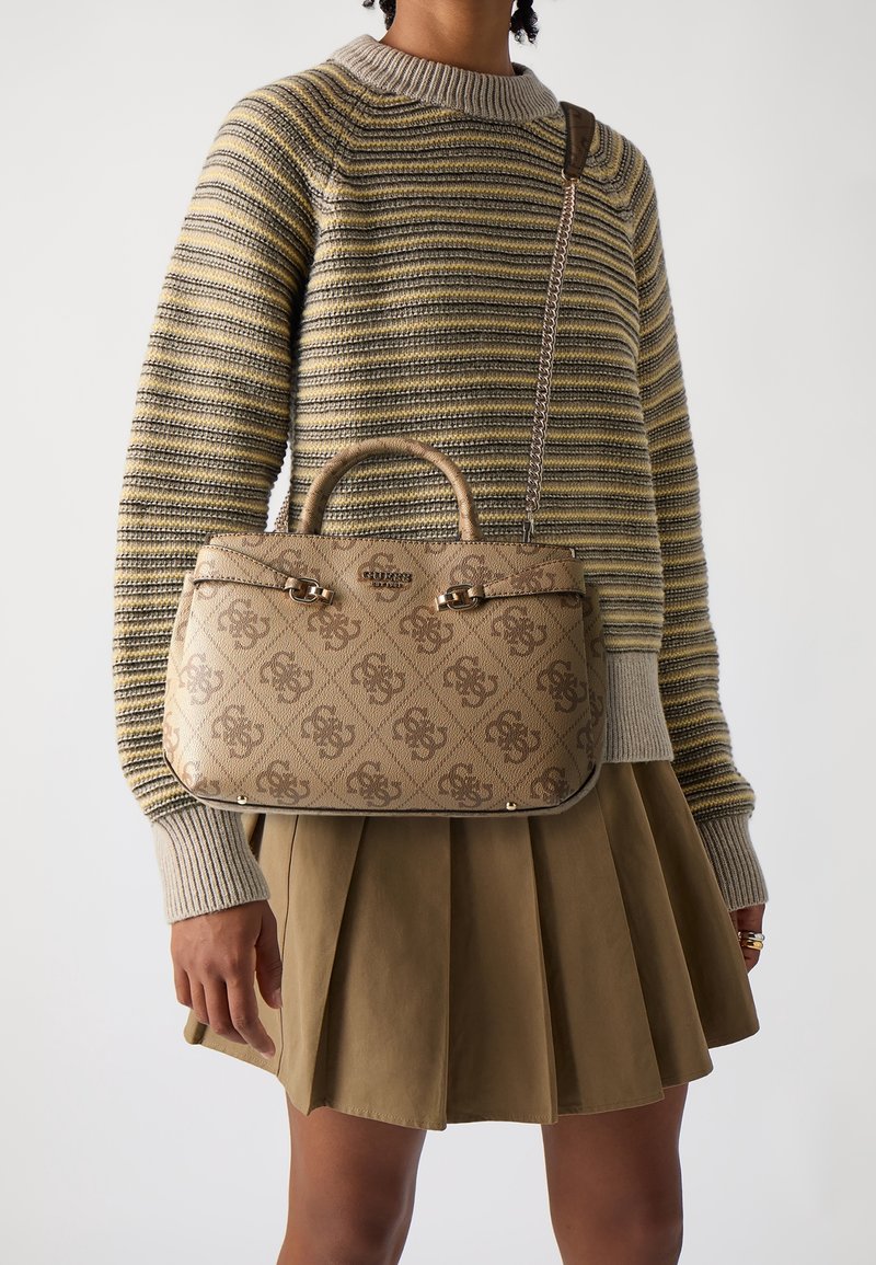 Personne portant une jupe plissée beige et un pull en maille rayé, tenant un sac à main marron clair à motifs avec poignées supérieures et bandoulière.