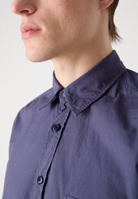 Menns mørk lilla knapp-up skjorte, med en klassisk krage, blå knapper, og et lett, teksturert stoff. Subtile detaljer på plaket.