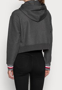 Sudadera gris recortada con capucha ajustable, dobladillo ribeteado y puños con rayas rojas y plateadas. Material suave con una textura lisa.