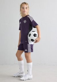 Lila Fußballuniform mit schwarzen Akzenten, weißen Streifen und dem Manchester United Logo. Weiße Socken und Fußballschuhe; hält einen schwarz-weißen Fußball.