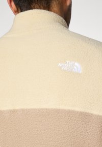 Fleece-jakke i krem og brunt, med høy krage, myk tekstur og hvit brodert logo på ryggen.